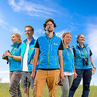 Gruppe aus drei Frauen und zwei Männern in blauen Westen auf Wiese vor blauem Himmel