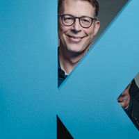 Staatsminister Markus Blume schaut hinter großem blauen Buchstaben K hervor